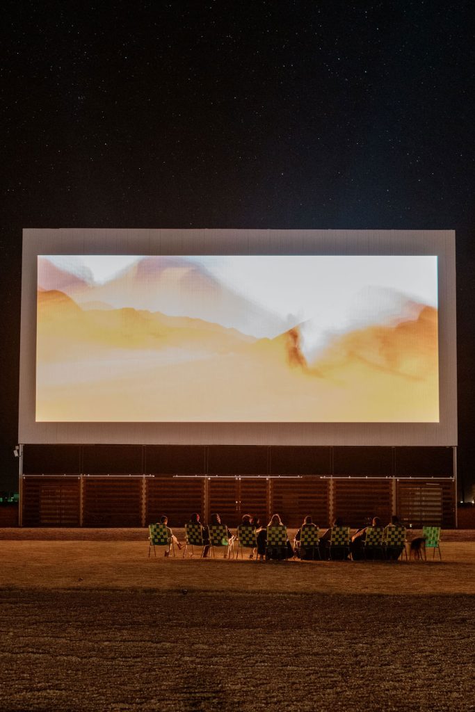 Group of people sitting in folder chairs on the lawn watching a movie on a big drive-in movie theater screen.