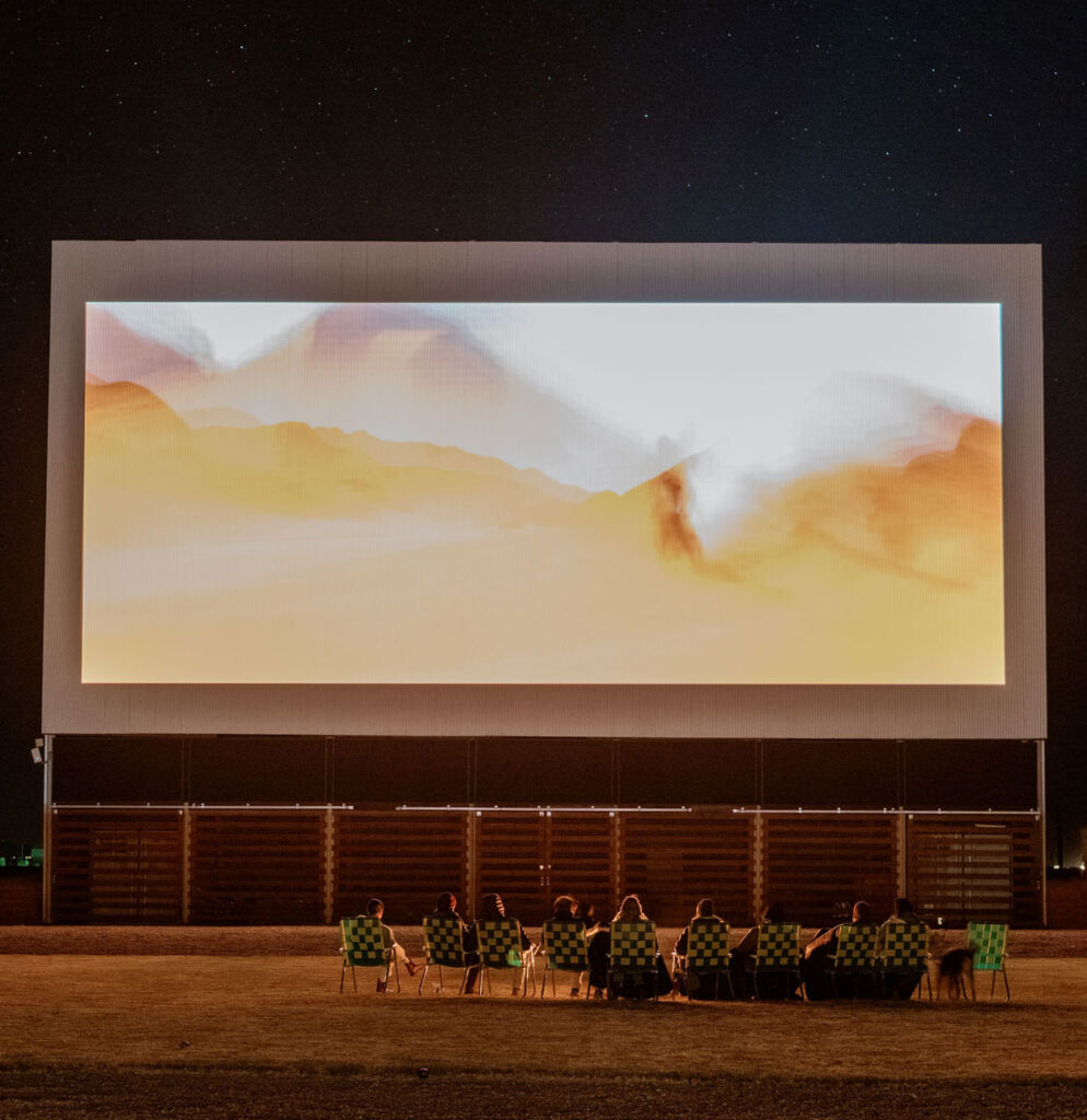 Group of people sitting in folder chairs on the lawn watching a movie on a big drive-in movie theater screen.